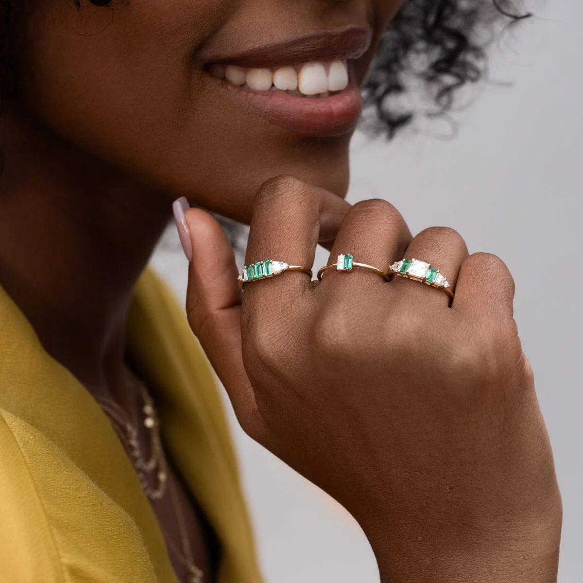 Close-up of a hand wearing 14k yellow gold Hall of Emerald Rings with natural rectangular emeralds and Asscher-cut diamonds