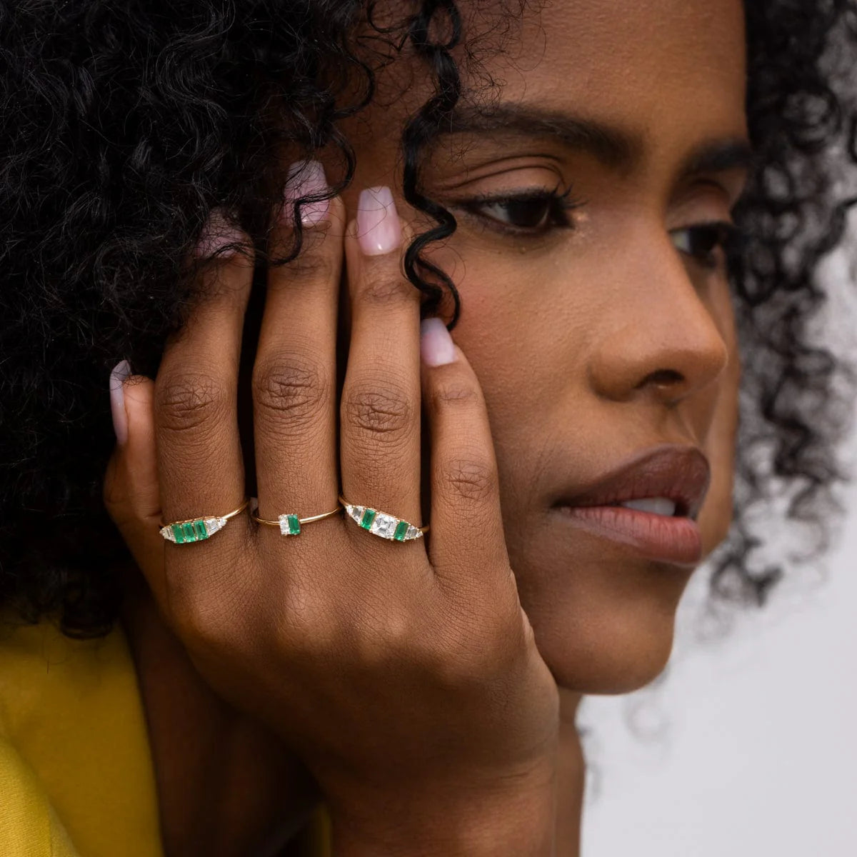 Close-up of a woman wearing 14k yellow gold Hall of Emerald Rings with natural rectangular emeralds and Asscher-cut diamonds on her fingers