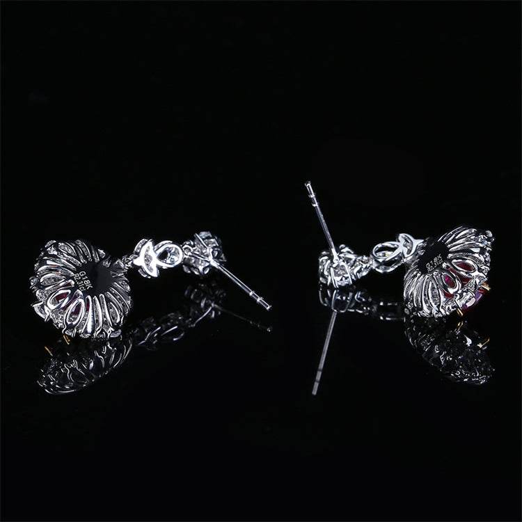 Detailed close-up of silver-toned drop earrings with intricate design and red ruby accents on black reflective surface